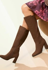 Botas marrones hasta la rodilla con un tacón grueso y cremallera lateral, combinadas con una falda floral, sobre un fondo suave y neutro.