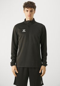 Jonge man die een zwarte sporttop met lange mouwen en rits en bijpassende korte broek draagt, staand tegen een effen lichte achtergrond.