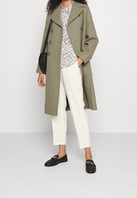 Manteau double boutonnage vert olive avec de grands boutons, associé à une chemise à fleurs, un pantalon blanc et des mocassins noirs avec des accents en chaîne.