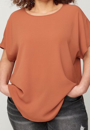 Personne portant un haut ample à manches courtes couleur rouille, les mains dans les poches d'un jean noir usé, vue du cou aux hanches.