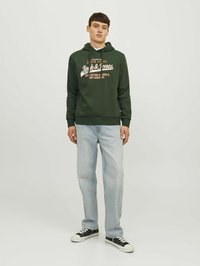 Sudadera verde con texto gráfico en blanco y naranja, combinada con jeans de un azul claro y zapatillas negras. Diseño simple con capucha ajustable.