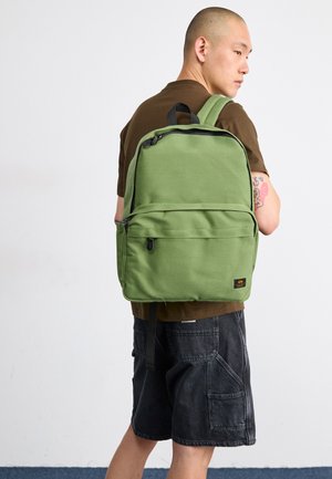 Green fabric backpack with a rectangular shape, front zip pocket, and black zipper accents. Worn on the back by a person in a brown shirt.