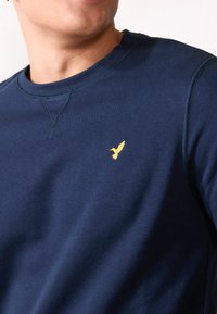 Navy blue sweatshirt with a round neckline, featuring a small gold bird logo on the left chest. Smooth texture and casual fit.