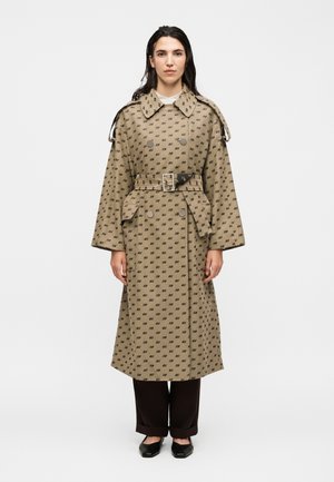 Femme debout portant un trench-coat beige ceinturé avec un motif noir "XX", pantalon noir et chaussures noires sur fond blanc.