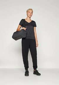 Fiatal személy rövid szőke hajjal áll, egy fekete sporttáska tartva, fekete pólót, fekete tréningnadrágot és fekete sportcipőt viselve egy sima háttér előtt.