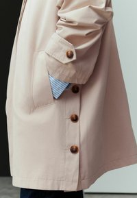 Hellrosa oversized Mantel mit braunen Knöpfen, Seitentaschen und hochgekrempelten Ärmeln, die ein blau-weiß gestreiftes Hemd darunter zeigen.