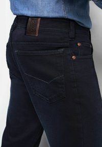 Dunkelblaue Jeans im geraden Schnitt, mit einem braunen Lederaufnäher an der Taille, Kupfennieten und einer einzelnen Gesäßtasche mit einem V-förmigen Design.