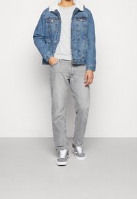 Jeansjacke mit Lammfellkragen, graues T-Shirt, hellgraue Jeans und graue Sneakers mit weißen Sohlen, die weiße Schnürsenkel und seitliche Akzente haben.