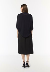 Schwarzes, halbtransparentes, übergroßes Oberteil mit abgerundetem Saum, kombiniert mit einem schwarzen, knielangen Rock, und akzentuiert durch hochhackige, offene Schuhe.