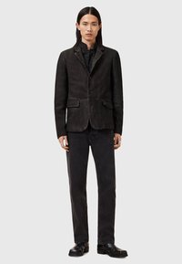 Blazer de cuero negro con solapa, cierre frontal de botones, dos bolsillos de solapa y un acabado ligeramente texturizado, combinado con jeans negros y botas.