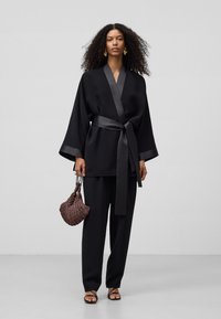 Zwarte kimono-stijl top met satijnen accenten, brede mouwen, vastgebonden in de taille, gecombineerd met losse zwarte trousers en een gestructureerde, bewerkte handtas.