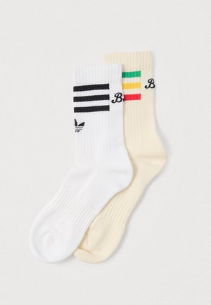 Two pairs of socks: one white with black stripes and branding, the other cream with colorful horizontal stripes and branding. Ribbed texture.