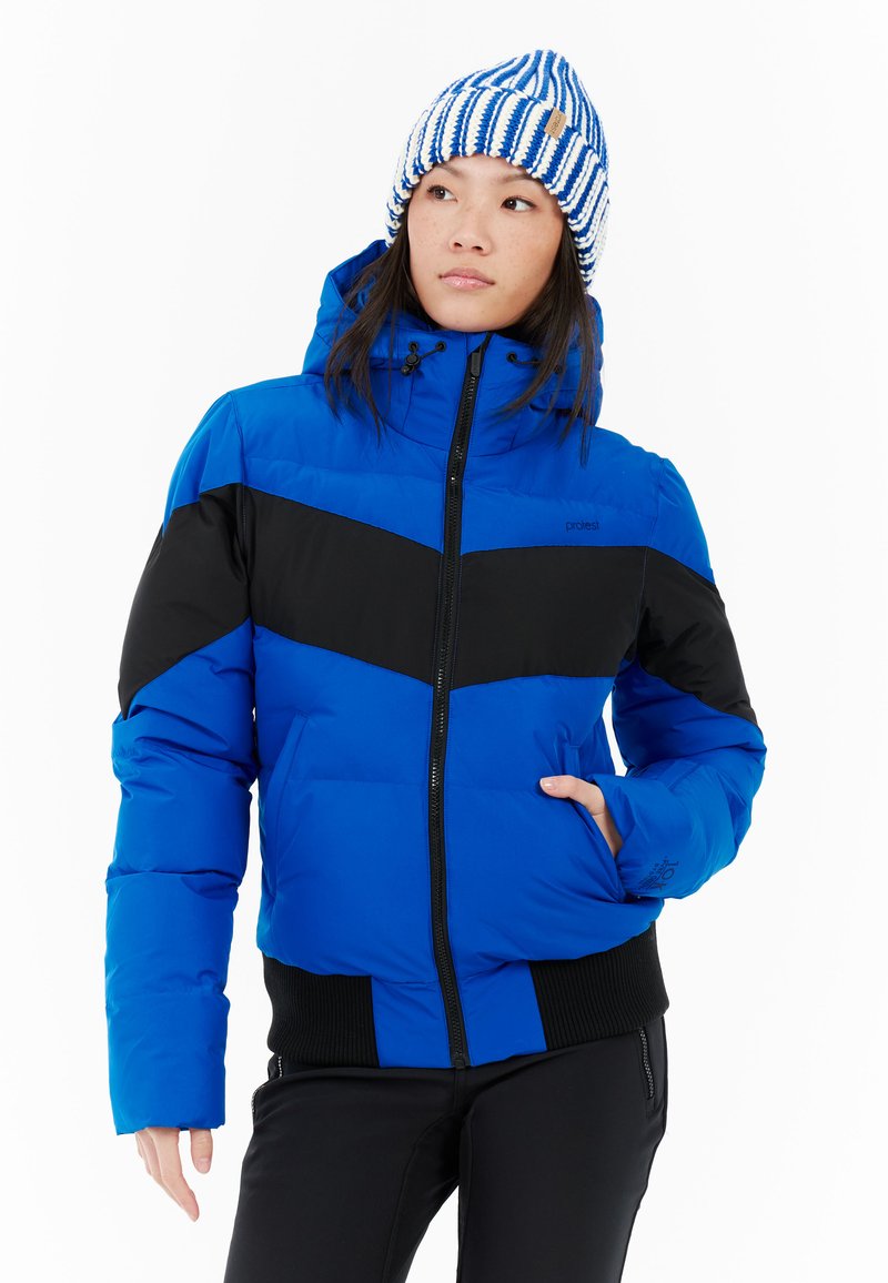 Žena v modrej a čiernej prešívanej bunde a modro-bielej pruhovanej pletenej čiapke, pózujúca s jednou rukou v vrecku na bielom pozadí.