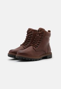 Botas de cuero marrón con un acabado suave, tobillo alto, punta redonda y suela de goma negra resistente. Atadas con siete ojetes metálicos y un lazo en la parte trasera.