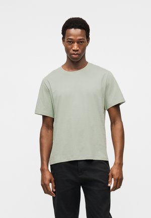 Žalios mėtos spalvos trumpomis rankovėmis medvilniniai marškinėliai su apvalia iškirpte, turintys laisvą siluetą ir minimalistinį dizainą. Dėvimi kartu su juodomis kelnėmis.