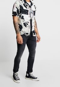 Košile s krátkým rukávem s květinovým a abstraktním vzorem v černé, krémové a růžové barvě, spárovaná s černými slim džíny a černými vysokými teniskami.