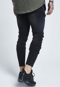 Svart slitna skinny jeans med en avsmalnande passform och synlig sömnad, i kombination med svarta sportskor som visar en texturerad yta.
