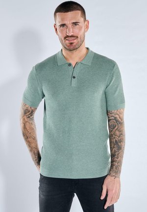 Mann mit kurzen Haaren und Tätowierungen auf beiden Armen, trägt ein hellgrünes, kurzärmeliges, gestricktes Poloshirt und dunkle Hosen vor grauem Hintergrund.