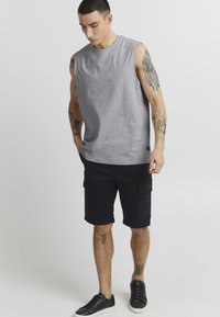 Graues ärmelloses T-Shirt aus weichem Stoff mit rundem Ausschnitt; kombiniert mit dunklen Cargo-Shorts mit seitlichen Taschen und schwarzen Turnschuhen.