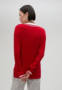 Red ribbed sweater with long sleeves, white striped accents at the collar and cuffs, paired with striped pants. Smooth texture.