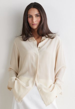 Kvinde med langt mørkt hår iført en løs, lysebeige skjorte med knapper og hænderne i lommerne mod en ensfarvet hvid baggrund.