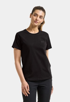 Kobieta z kucykiem, ubrana w prostą czarną koszulkę z krótkim rękawem i czarne spodnie, stoi z prawą ręką luźno przy boku na białym tle.