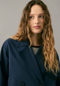 Abrigo azul marino cruzado con cuello de muesca, confeccionado en una tela lisa, combinado con un suéter gris debajo.