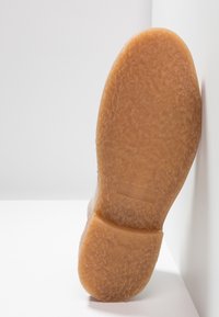 Semelle d'une chaussure beige à semelle crêpe avec un motif texturé, photographiée sur fond blanc.