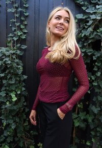 Bordowa bluzka z długim rękawem wykonana z prześwitującego materiału, ozdobiona małymi srebrnymi ćwiekami. Posiada dopasowany krój i ozdobne mankiety. Widoczne czarne spodnie.