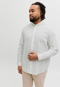 Homme avec barbe et dreadlocks portant une chemise blanche boutonnée et un pantalon beige, regardant sur le côté avec une main près de sa poitrine.