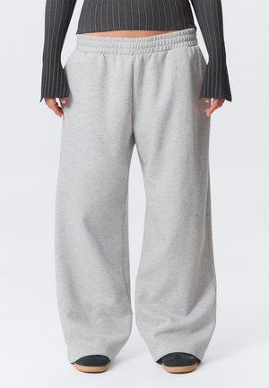 Personne portant un pantalon de survêtement ample gris clair avec taille élastique et des chaussures noires avec semelles brun clair, debout devant un fond uni.
