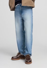 Jeans in denim azzurro chiaro con una vestibilità comoda, caratterizzati da una finitura sbiadita e design standard a cinque tasche, abbinati a scarpe marroni con lacci.