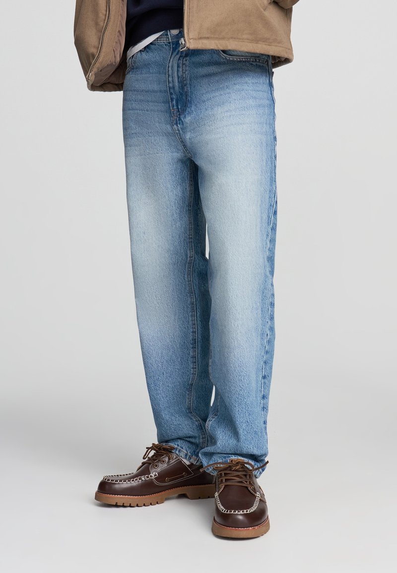 Jeans in denim azzurro chiaro con una vestibilità comoda, caratterizzati da una finitura sbiadita e design standard a cinque tasche, abbinati a scarpe marroni con lacci.