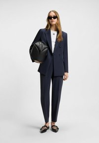 Tummansininen räätälöity puku, jossa on kaksirivinen bleiseri, valkoinen paita ja mustat loafer-kengät. Pitelee teksturoitua mustaa laukku, jossa on kartion muoto.