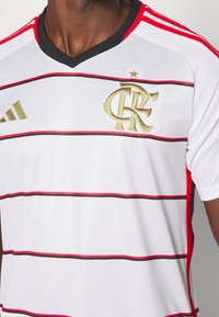 White sports jersey with black and red horizontal stripes. Features a black V-neck, gold embroidered logo, and adidas branding in gold.