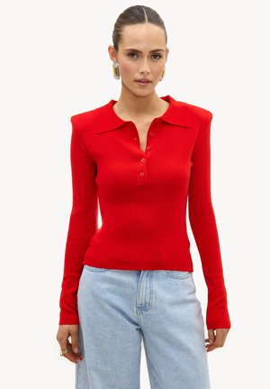 Woman with slicked-back hair wearing large silver earrings, a red long-sleeve buttoned top, and light blue jeans against a white background.