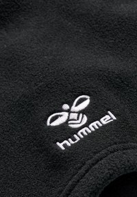 Czarny materiał polarowy o gładkiej strukturze. Posiada białe wyszywane logo pszczoły oraz napis "hummel". Widoczne zaokrąglone krawędzie.