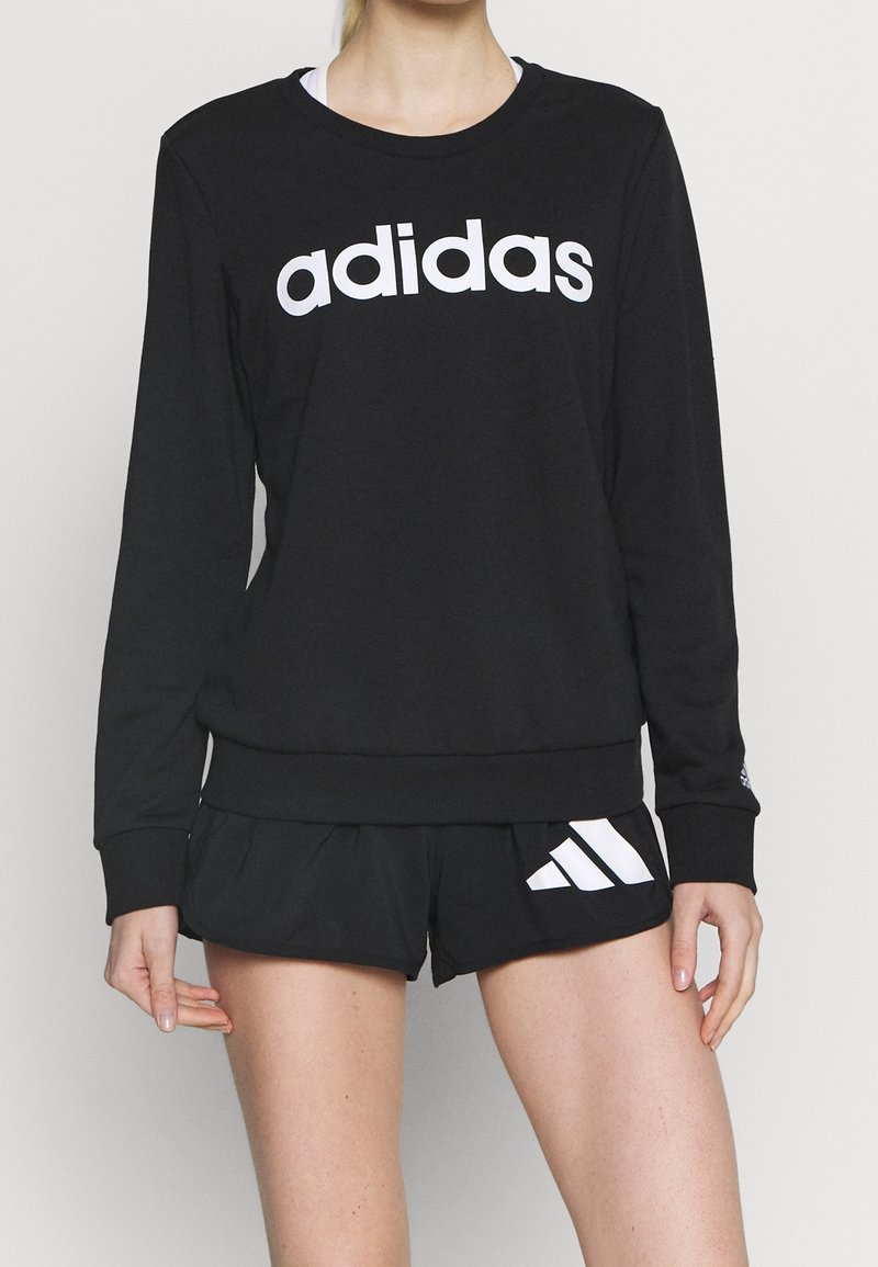 Personne portant un sweat-shirt Adidas noir avec un logo blanc et un short Adidas noir avec trois bandes blanches sur un fond clair uni.