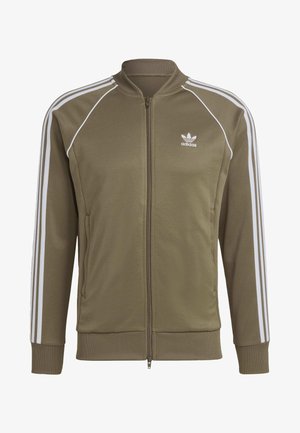 Homme portant une veste zippée Adidas vert olive avec des rayures blanches sur les manches et un jean bleu clair, main dans la poche de la veste.