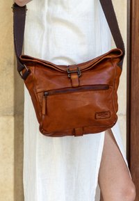 Brown leather shoulder bag with a zippered front pocket, adjustable fabric strap, and a fold-over top. Textured finish with subtle branding.