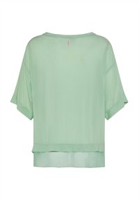 Mintgrøn kortærmet bluse med løs pasform og lagdelt kant, vist bagfra med en lille lyserød mærkat fra mærket nær halsudskæringen.
