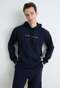 Hoodie bleu marine en tissu doux avec une poche avant et le logo "TOMMY HILFIGER" en lettres blanches et rouges sur la poitrine.