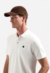 Bruine wollen pet met gebogen klep, met daarop een hexagonaal zwart logo op een wit poloshirt. Close-up zijaanzicht, neutrale achtergrond.