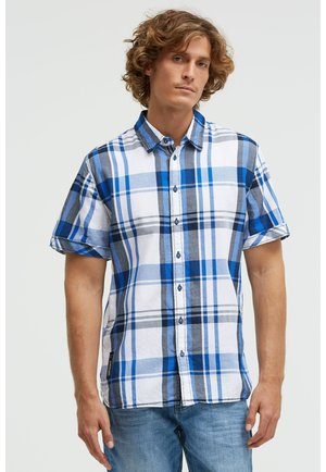 Junger Mann trägt ein kurzärmliges Hemd mit blau-weiß-grau kariertem Muster und hellblaue Jeans vor einem schlichten Hintergrund.