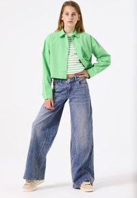 Grünes, gekürztes Hemd mit Knöpfen über einem gestreiften, hellgrünen und weißen Oberteil, kombiniert mit weitem blauen Jeans und beigen Sneakers.