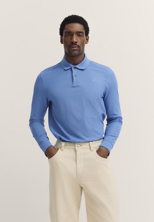 Himmelblaues langärmliges Poloshirt mit klassischem Kragen und drei Knöpfen, kombiniert mit hellcremefarbenen Hosen, die über seitliche Taschen verfügen.