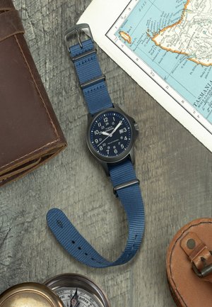 Reloj con correa de nylon azul, caja negra y esfera azul, que presenta números blancos, función de fecha y una correa texturizada, colocado sobre madera.