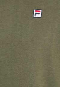 T-shirt verde oliva in morbido cotone, con un piccolo logo ricamato in rosso, bianco e blu sulla parte superiore sinistra.