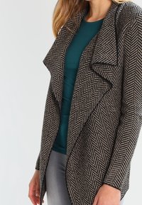Cappotto con motivo pied de poule in beige e nero, caratterizzato da un colletto drappeggiato e apertura frontale, abbinato a una maglia teal e pantaloni grigi.