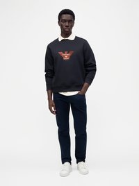 Мъж, облечен в черна суитшърт Emporio Armani, бяла риза с яка отдолу, тъмни панталони и бели маратонки, стоящ с една ръка в джоба.